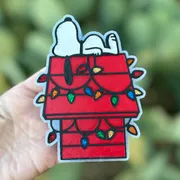 Christmas car freshie shaped like a sleeping cartoon dog on a red doghouse draped with colorful glitter Christmas lights in a stained-glass style, held in hand against greenery.