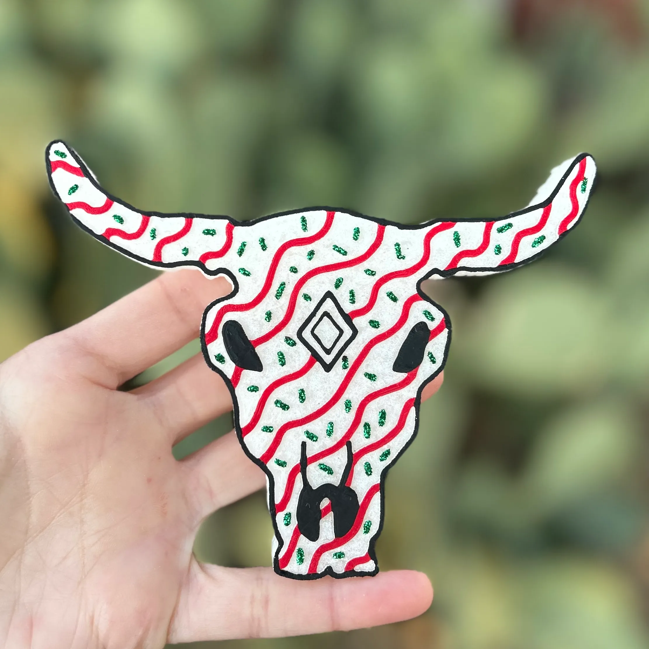 Hand-painted white longhorn steer skull car freshie decorated with red wavy stripes and small green holiday sprigs, held in a hand in front of a green cactus background, in a western Christmas style.