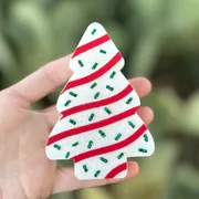 Christmas tree shaped car freshie made to look like a snack cake with white frosting, red icing stripes, and green glitter sprinkles, held in hand against a green background.