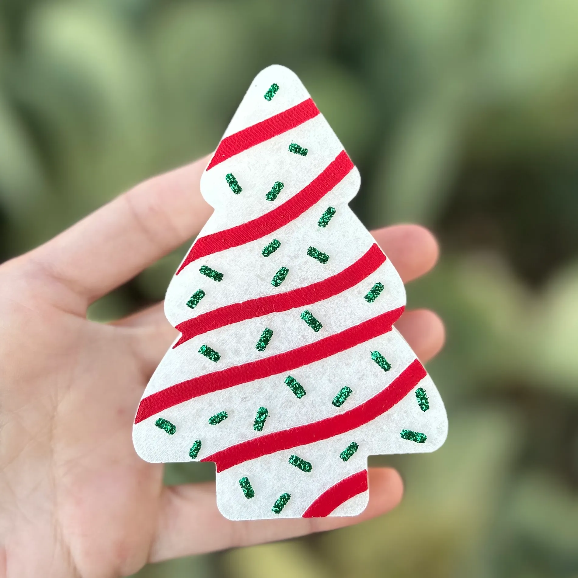Christmas tree shaped car freshie made to look like a snack cake with white frosting, red icing stripes, and green glitter sprinkles, held in hand against a green background.