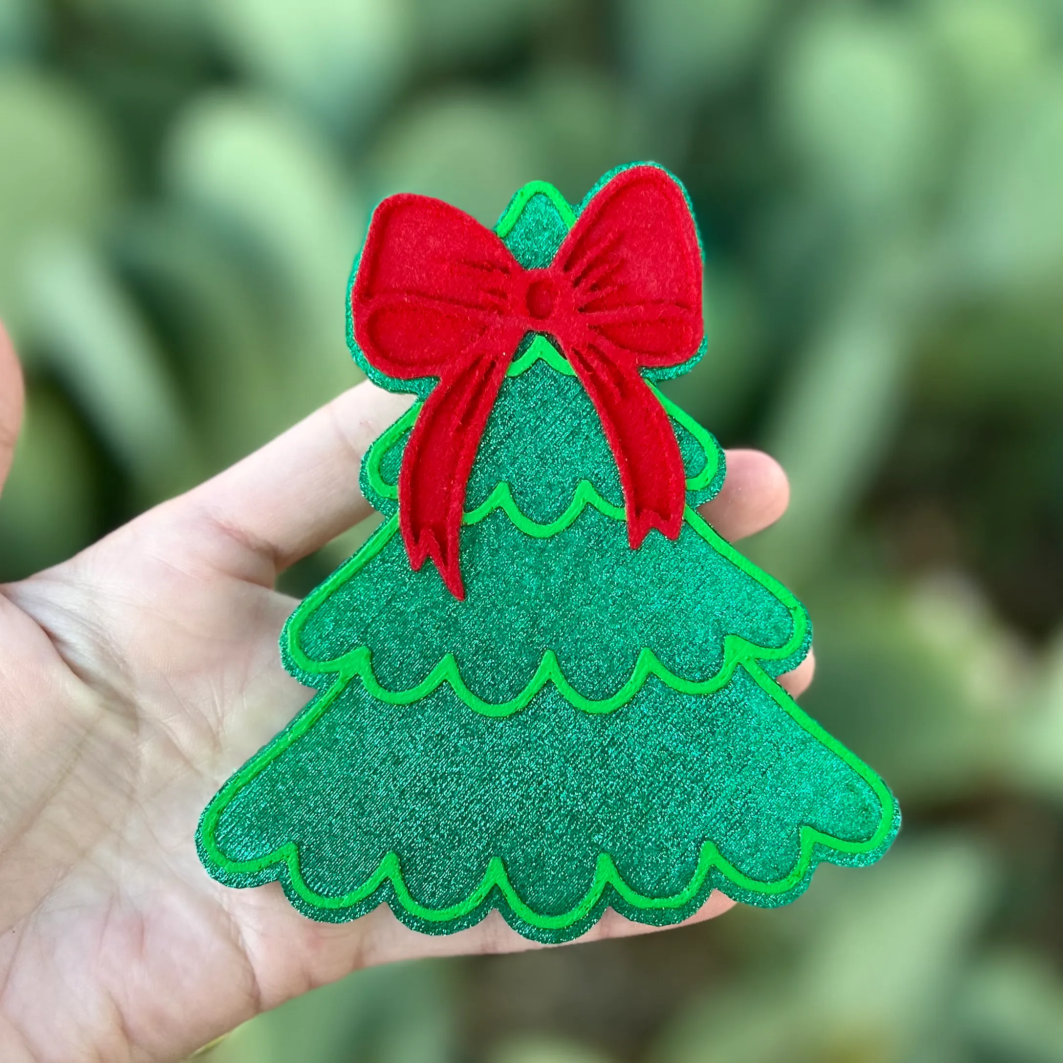 Green glitter Christmas tree car freshie with scalloped branches and a red bow on top, held in a hand against a cactus background.