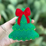 Green glitter Christmas tree car freshie with scalloped branches and a red bow on top, held in a hand against a cactus background.