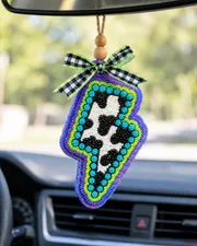 Handmade lightning bolt shaped car freshie in black and white cow print, a scented aroma bead air freshener hanging from a car rearview mirror.