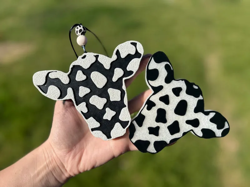 Handmade scented aroma bead car freshie shaped like a black-and-white cowhide steer head with ears up on a cream base, a western farmhouse air freshener on a cow-print beaded cord.