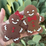 Two handmade gingerbread car freshies held in a hand, a gingerbread girl with a red glitter bow and a gingerbread boy with a red bow tie, both with white icing details, against a green cactus background.
