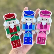 Three glitter Nutcracker Soldier car freshies held in a hand, in red and green, royal blue, and pink color variations, each a scented Christmas car air freshener.