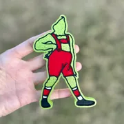 Handmade Christmas car freshie shaped like a green grump character in red overalls, held in hand over grass, a scented aroma bead car air freshener.