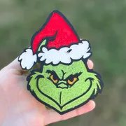 Handmade Christmas car freshie shaped like a grumpy green character wearing a red and white Santa hat, scented aroma bead air freshener held in hand outdoors.