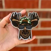 Handmade scented aroma bead car freshie air freshener shaped like a cartoon buck deer with big antlers wearing black sunglasses, a camo backwards cap, and a brown beard, hanging from a rearview mirror.
