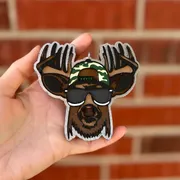 Handmade scented aroma bead car freshie air freshener shaped like a cartoon buck deer with big antlers wearing black sunglasses, a camo backwards cap, and a brown beard, hanging from a rearview mirror.