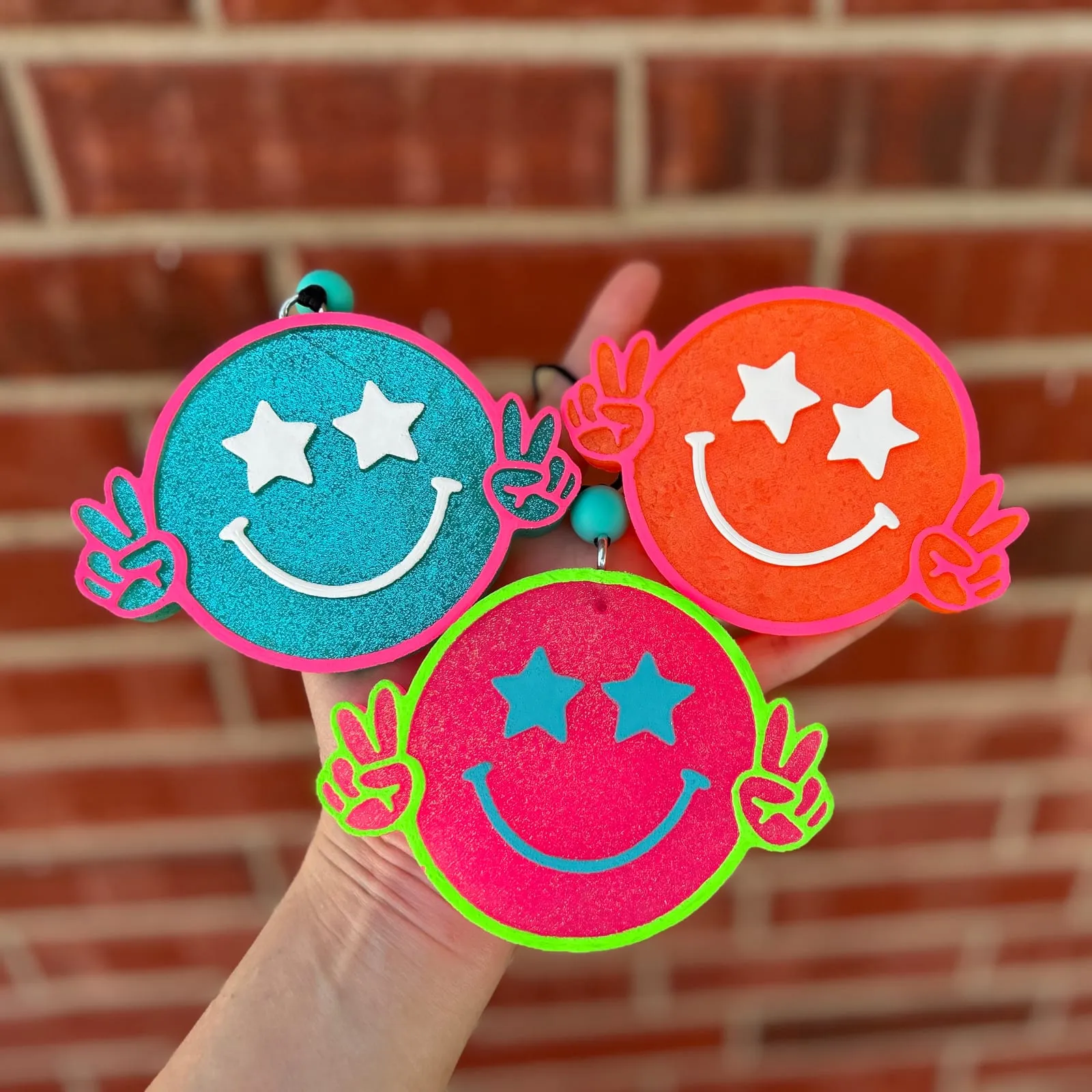 Handmade scented aroma bead car freshie air freshener shaped like a round retro smiley face with puffy star eyes and little hands throwing peace signs, hanging from a beaded cord.
