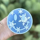Round blue car freshie shaped like a smiley face decorated with white snowflakes, held in hand against a green plant background.